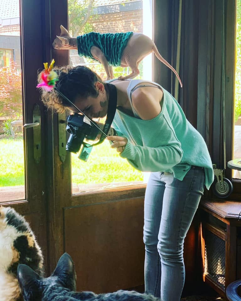 Femme qui prend une photo penchée en avant avec un chat sphynx qui lui monte sur le dos avec un pull vert