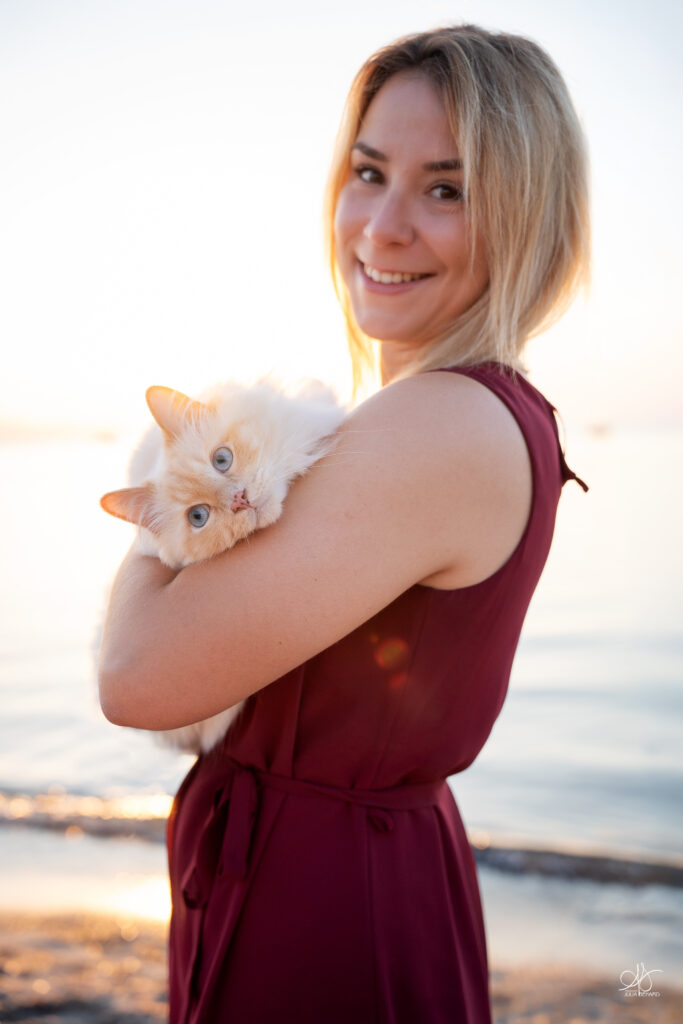 Jeune femme blonde en bord de mer avec son chat type sacré de birmanie aux bras, elle porte une robe rouge